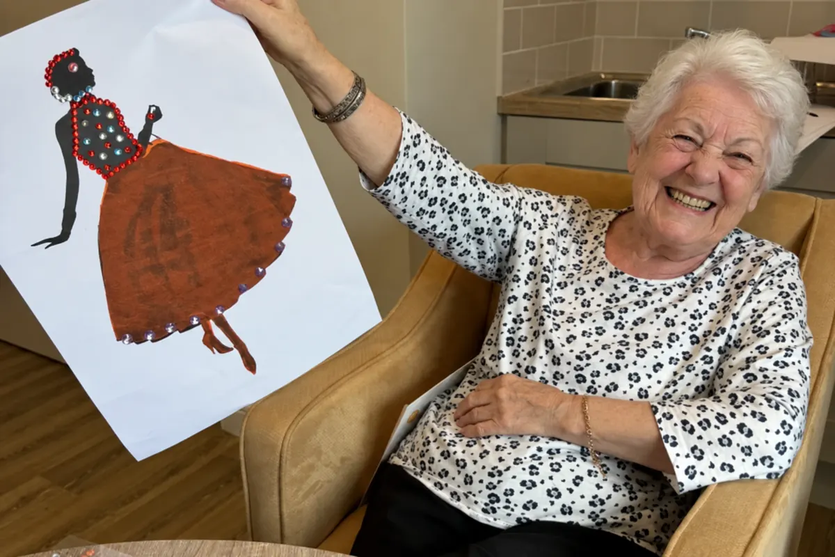 A resident in an armchair laughing as she holds up her finished picture of a dancer in a jewelled dress
