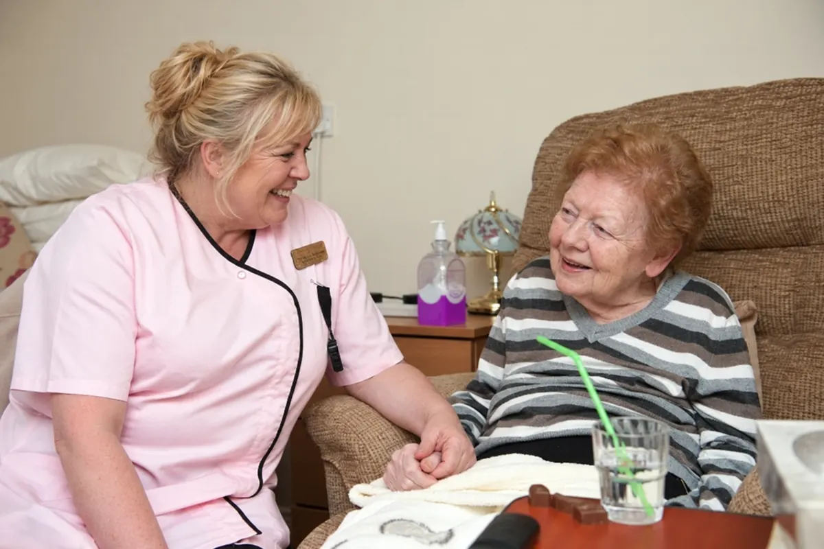A carer in a pink tunic holding a resident’s hand as they smile at each other beside her armchair