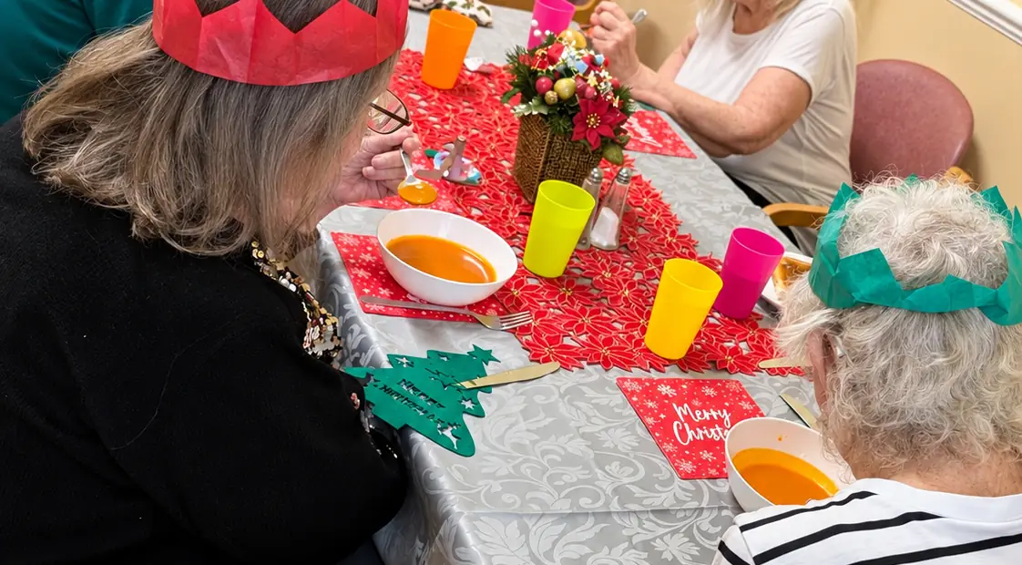 Christmas lunch at Aquarius Lodge: residents in red and green paper crowns at a table laid with a red runner, Christmas mats and bowls of soup