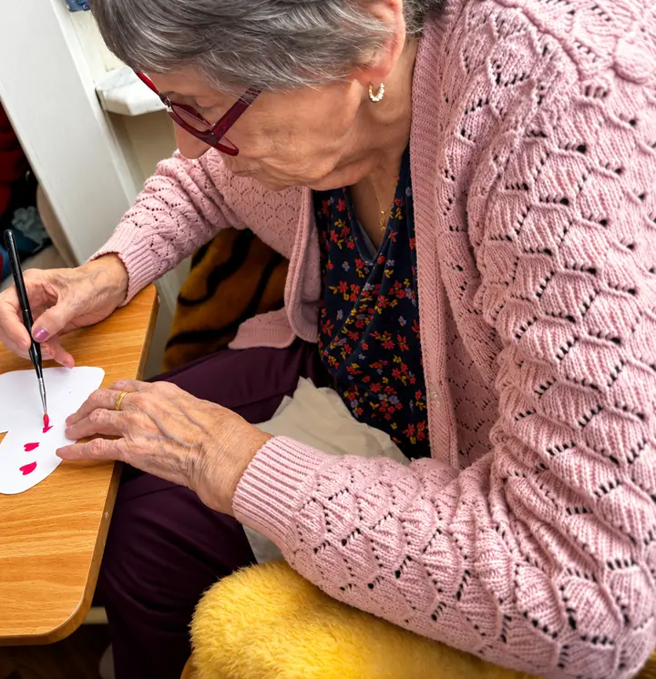 A resident in a pink cardigan and red glasses painting small red hearts on a paper heart