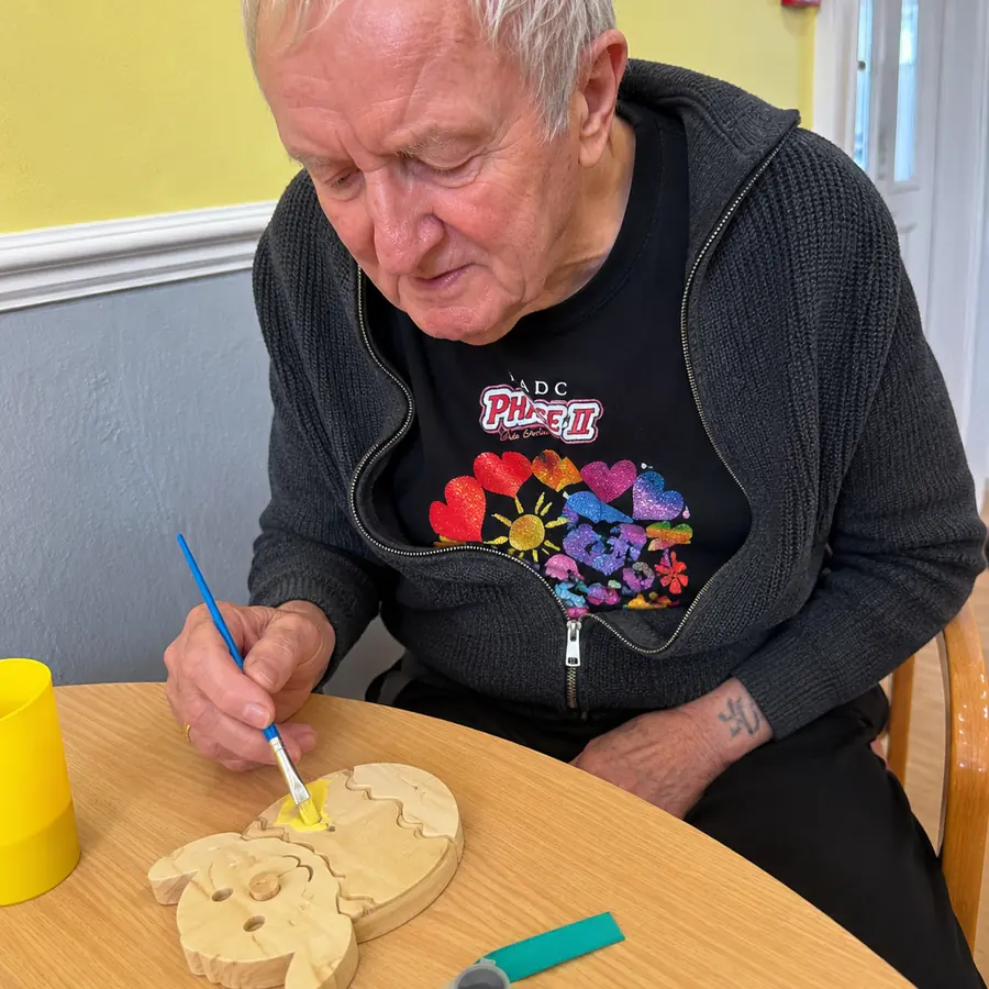 A resident in a dark cardigan painting a wooden cut-out yellow at the table